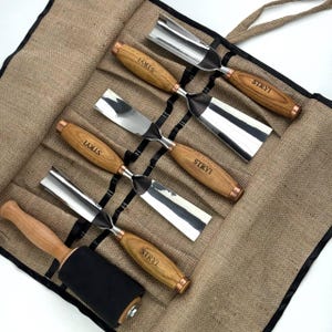 May include: A set of five wood carving tools and a mallet with black heads and light brown wooden handles, each branded with the text STRYI. The tools are arranged in a tan burlap roll-up organizer with individual pockets for each item.