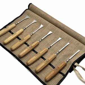 May include: A set of six metal wood carving tools with light brown wooden handles, each engraved with the brand name STRYI. The tools are arranged in a tan fabric roll-up organizer with black trim and individual slots for each tool.