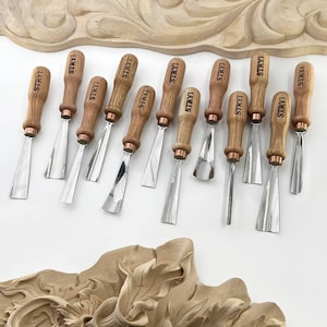 May include: A collection of wood carving chisels with wooden handles and silver blades. Each chisel is marked with the word "STRYI" on the handle. The tools are arranged on a white surface, with a carved wooden decorative element in the background.