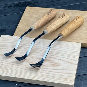 May include: Three wood carving tools with polished metal curved blades and light brown wooden handles. Each handle is engraved with the brand name STRYI in black lettering. The tools feature a distinct bent shank design for detailed woodworking.