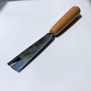 以下が含まれることがあります： 木製のハンドルが付いた金属製の彫刻ツールで、先端は斜めにカットされています。ハンドルは明るい色の木でできており、"S.L.S."という文字が刻まれています。