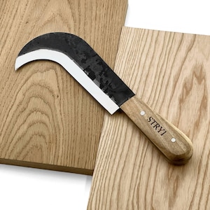 May include: A curved, black blade knife with a wooden handle. The blade has a white edge and the handle is engraved with "STRYI". The knife is displayed on a light-coloured wooden surface.