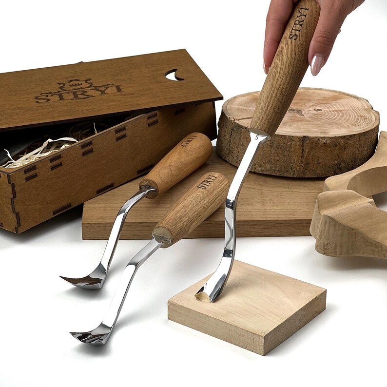 May include: Three metal wood carving tools with light brown wooden handles, each branded with the STRYI logo. The set includes a wooden storage box with the STRYI logo, a circular wood slice, and a square wooden block for tool testing.