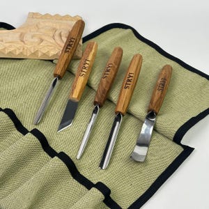 Puede incluir: Conjunto de cinco herramientas de talla de madera con mangos de madera. Las herramientas están dispuestas en un rollo de tela verde con ribete negro. Cada mango está grabado con la palabra "STRYI".