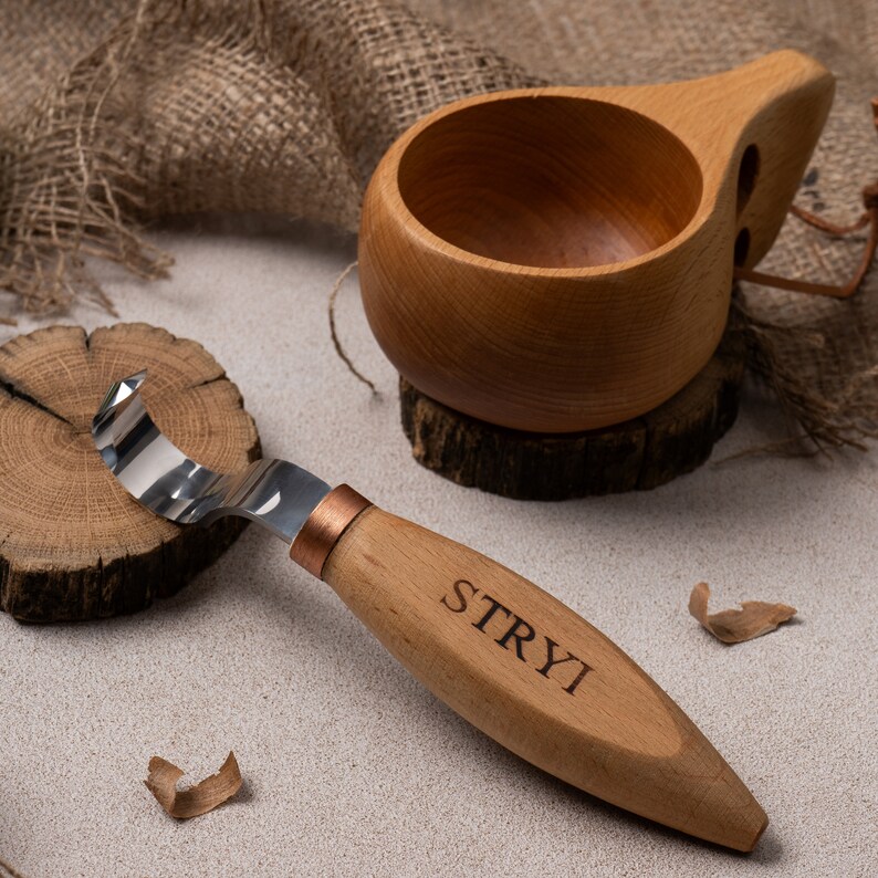 May include: A wooden carving knife with a copper-colored ferrule and a wooden handle engraved with the word "STRYI". The knife is resting on a wooden disc. A wooden cup with a handle is in the background.