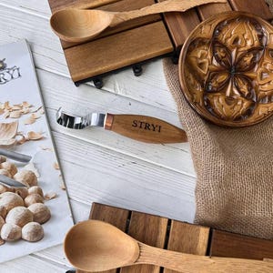 May include: A wooden spoon, a wooden carving tool with a metal blade, and a wooden plate with a carved design. The carving tool has the word "STRYI" engraved on the handle.