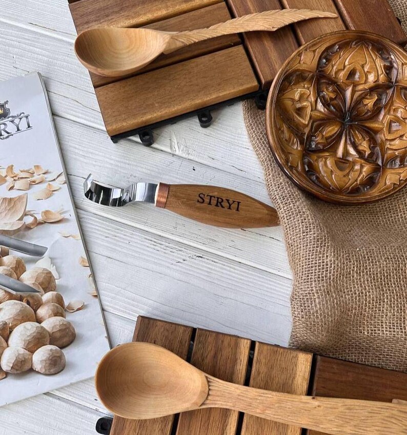 May include: A wooden spoon, a wooden carving tool with a metal blade, and a wooden plate with a carved design. The carving tool has the word "STRYI" engraved on the handle.