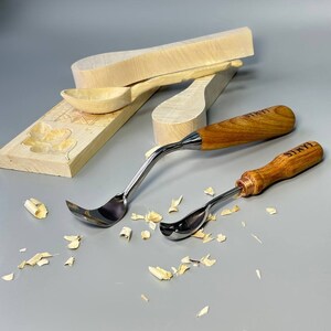 May include: Two wood carving gouges with curved stainless steel blades and wooden handles. The handles are engraved with the word "STRYI". Several pieces of wood shavings are scattered around the tools, along with unfinished wooden spoon blanks.