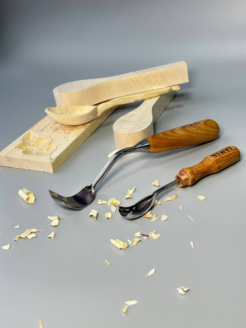 May include: Two wood carving gouges with curved stainless steel blades and wooden handles. The handles are engraved with the word "STRYI". Several pieces of wood shavings are scattered around the tools, along with unfinished wooden spoon blanks.