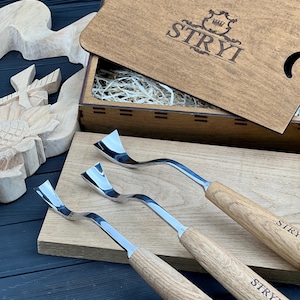 Könnte beinhalten: Drei Holzschnitzwerkzeuge aus Metall mit gebogenen Klingen und hellbraunen Holzgriffen, jeweils mit dem Text STRYI versehen. Die Werkzeuge liegen auf einem Holzbrett neben einer Holzkiste mit dem STRYI-Logo und einer geschnitzten Holzsonnenblume.