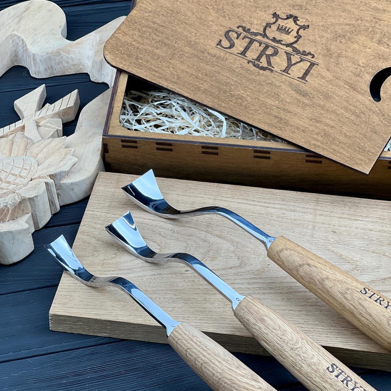 May include: Three metal wood carving tools with curved blades and light brown wooden handles, each stamped with the text STRYI. The tools are arranged on a wooden board next to a wooden box with the STRYI logo and a carved wooden sunflower.