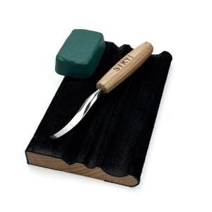 May include: A wood carving tool with a curved metal blade and a light wood handle featuring the text STRYI, accompanied by a rectangular green polishing compound block and a black leather strop mounted on a wooden base with grooved channels.