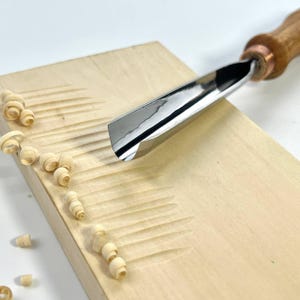 Puede incluir: Una gubia de carpintería con una hoja de acero pulido y un mango de madera está tallando un bloque de madera de color claro. Las virutas de madera están esparcidas alrededor de las ranuras talladas, mostrando el uso de la herramienta para dar forma a la madera. La gubia está en ángulo, lista para hacer otro corte.