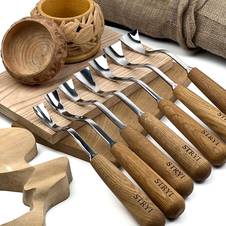 Peut inclure: Un ensemble de six gouges &agrave; bois en m&eacute;tal avec des lames incurv&eacute;es et des manches en bois brun clair, chacun marqu&eacute; du texte STRYI. Les outils sont dispos&eacute;s sur une planche en bois &agrave; c&ocirc;t&eacute; d'un bol en bois sculpt&eacute; et d'un bloc en bois.