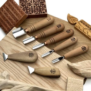 May include: A set of six wood carving tools with light brown wooden handles, each featuring a small stamped logo. The collection includes four gouges with curved metal blades and two knives with triangular metal blades, arranged on a wood surface.