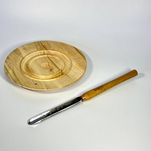 Puede incluir: Un recipiente de mantequilla de madera con una tapa redonda y un pequeño cuchillo de mantequilla de madera con una hoja de metal. El cuchillo tiene la palabra "STRYJ" grabada en el mango.