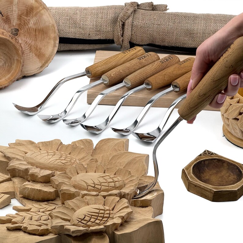Peut inclure: Un ensemble de six outils de sculpture sur bois avec des lames en m&eacute;tal poli et des manches en bois brun clair, chacun portant la marque STRYI grav&eacute;e. Les outils sont align&eacute;s, l'un d'eux &eacute;tant utilis&eacute; pour sculpter un motif de tournesol.