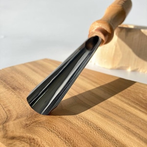 以下が含まれることがあります： 木製のハンドルと金属製の刃を持つ木彫りの道具のクローズアップ。刃は湾曲しており、鋭い刃を持っています。道具は木の板の上に置かれています。