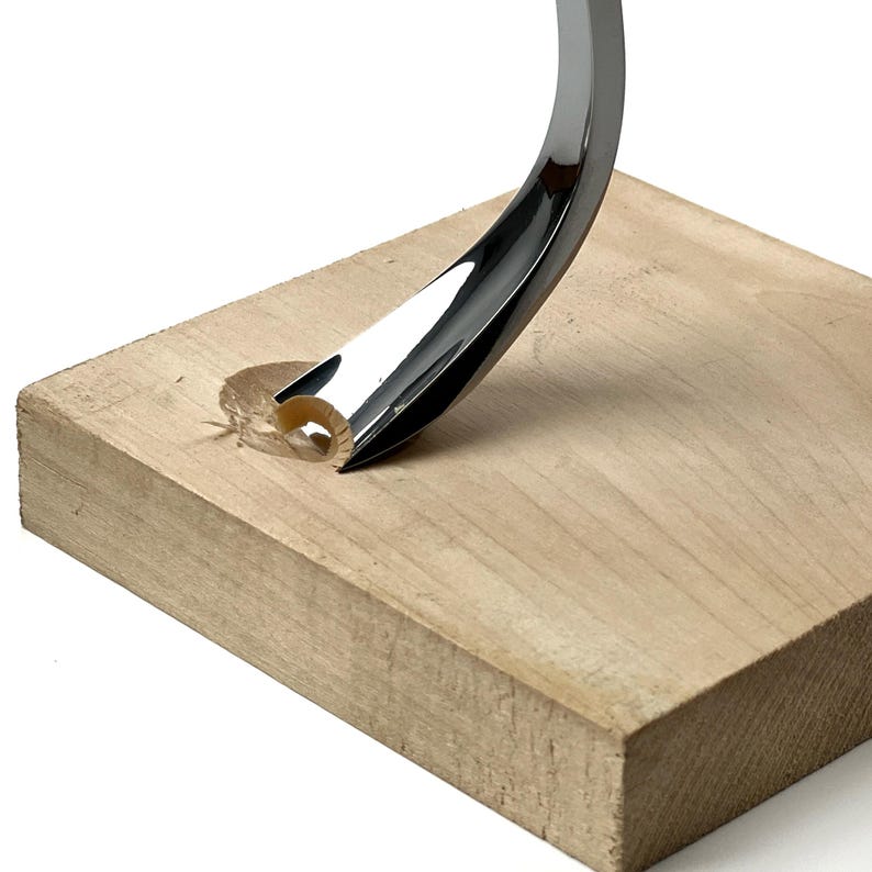 May include: A curved, polished chrome metal tool with a sharp, gouge-like tip is embedded into a square block of light-coloured, unfinished wood. The metal tool features a reflective, silver-toned surface and a concave, semi-circular cutting edge.