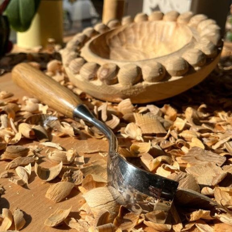 以下が含まれることがあります： 装飾的な縁が付いた木製のボウルと、木製の柄と曲がった金属製の刃が付いた彫刻ツール。 ボウルとツールは木くずで囲まれており、木工プロジェクトを示唆しています。
