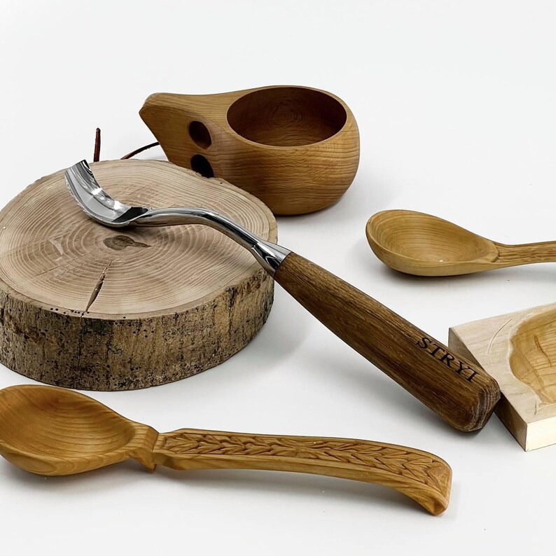 May include: A metal spoon with a curved neck and a wooden handle engraved with the text STRYT, positioned alongside a wooden cup, a plain wooden spoon, and a wooden spoon with a carved leaf pattern on the handle, resting on a round wood slice.