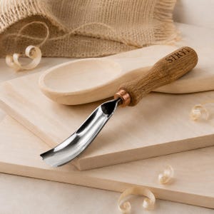 May include: A metal wood carving gouge with a curved, polished steel blade and a light brown wooden handle featuring the brand name STRYI engraved on the side. The tool is positioned on a light-colored wooden surface with wood shavings.