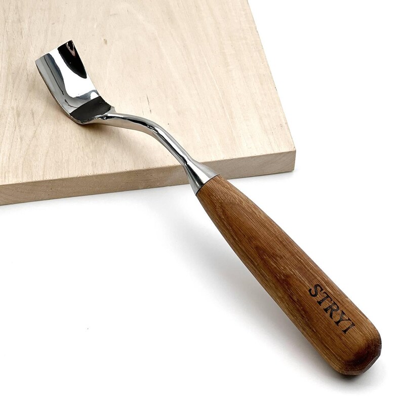 May include: A wood carving tool with a curved, polished metal blade and a smooth, light brown wooden handle. The word STRYI is clearly printed in black ink on the side of the handle, which features a visible wood grain pattern throughout.