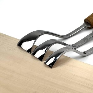 May include: Three metal wood carving gouges with curved, polished steel blades and wooden handles. The tools are arranged side-by-side, showing their distinct, sharp, concave edges designed for shaping wood, resting against a light wood surface.