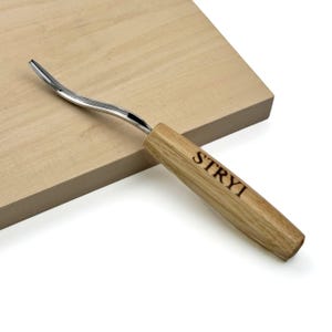 Puede incluir: Una herramienta de tallado en madera de metal con una hoja curva y un mango de madera marrón claro. El mango presenta la marca STRYI grabada en la madera. La herramienta está colocada en diagonal sobre una superficie de bloque de madera clara.