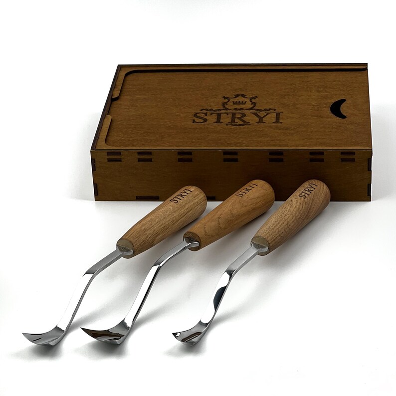 May include: Three metal carving tools with light brown wooden handles, each featuring the brand name STRYI engraved on the handle. The tools are positioned in front of a rectangular wooden storage box with the STRYI logo and a crescent moon icon.