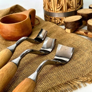 以下が含まれることがあります： 木製のハンドルと湾曲した金属製の刃が付いた木工工具のクローズアップ。ハンドル付きの木製カップと彫刻された木製のバケツも表示されます。ツールはジュートの表面に配置されています。