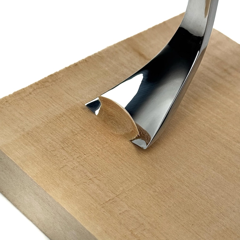 Peut inclure: Un outil de gouge en m&eacute;tal incurv&eacute; avec une finition argent&eacute;e polie et r&eacute;fl&eacute;chissante est montr&eacute; en train de sculpter un bloc de bois brun clair. La lame tranchante en forme de U est enfonc&eacute;e dans le bois, cr&eacute;ant une rainure semi-circulaire.