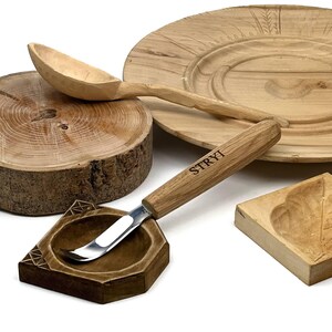 May include: A wood carving tool with a metal hook blade and a light brown wooden handle featuring the text STRYI, positioned near a carved wooden spoon, a circular wooden base, a hexagonal wooden block, and a square wooden piece with a leaf design.
