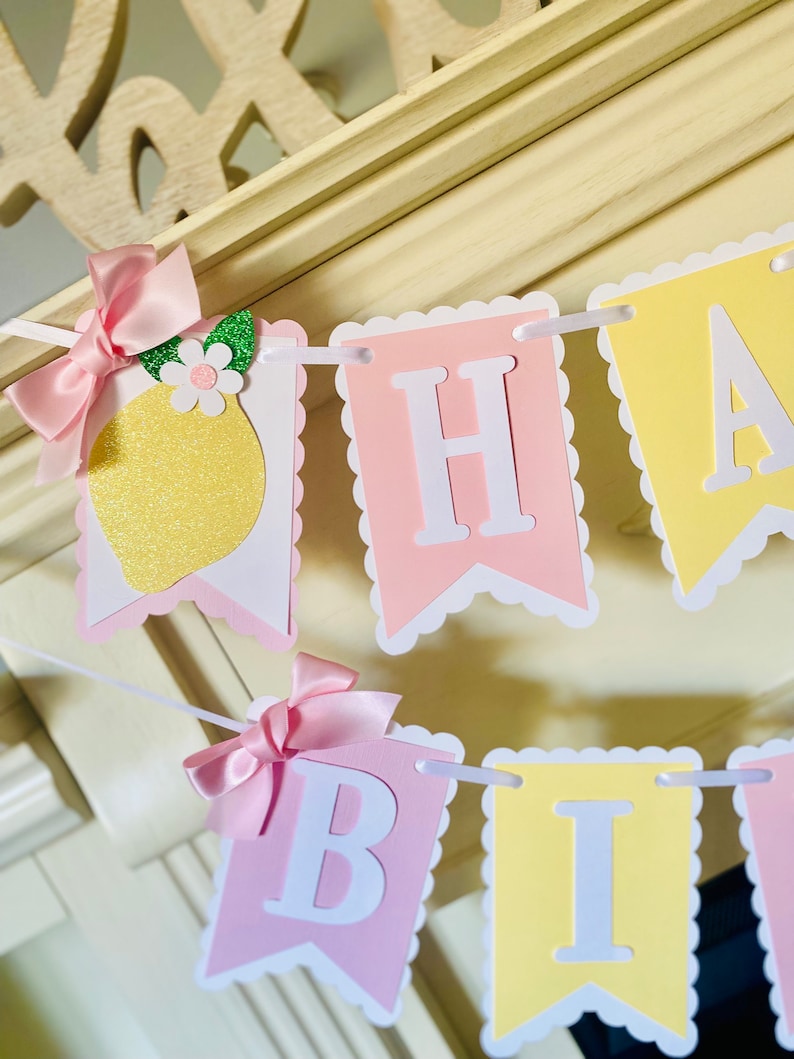 May include: A pink and yellow banner with scalloped edges that reads "Happy Birthday". The banner is decorated with a lemon, a pink bow, and a green leaf.