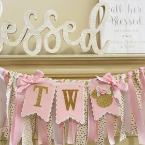 May include: A pink and gold banner with the letters "TW" and a Minnie Mouse silhouette. The banner has pink bows and ribbons, with white and gold polka dot fabric. A wooden sign with the word "Blessed" is in the background.