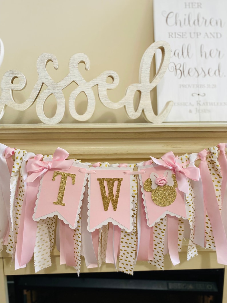 May include: A pink and gold banner with the letters "TW" and a Minnie Mouse silhouette. The banner has pink bows and ribbons, with white and gold polka dot fabric. A wooden sign with the word "Blessed" is in the background.