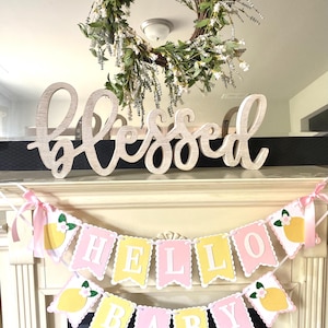 May include: A decorative display featuring a wooden "Blessed" sign, a floral wreath, and a banner that reads "Hello Baby." The banner has pink and yellow pennants with lemon and flower accents, perfect for a baby shower.