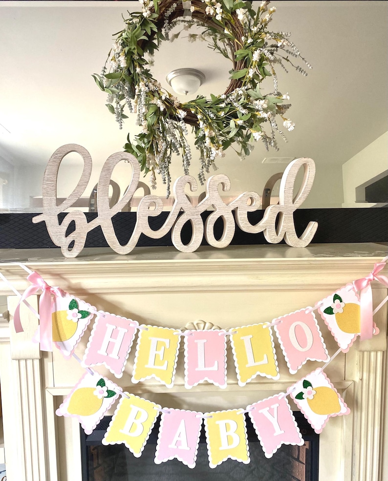 May include: A decorative display featuring a wooden "Blessed" sign, a floral wreath, and a banner that reads "Hello Baby." The banner has pink and yellow pennants with lemon and flower accents, perfect for a baby shower.