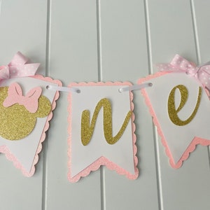 Puede incluir: Pancarta de "uno" con purpurina rosa y dorada con una cabeza de Minnie Mouse de purpurina rosa y dorada en el lado izquierdo. La pancarta est&aacute; enhebrada con una cinta blanca y tiene bordes festoneados rosas.