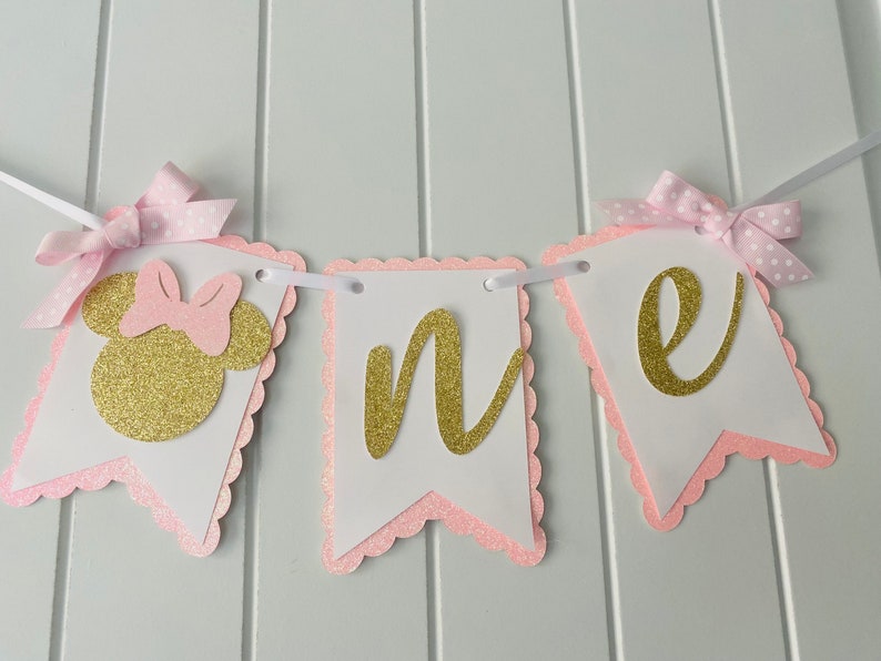 Puede incluir: Pancarta de "uno" con purpurina rosa y dorada con una cabeza de Minnie Mouse de purpurina rosa y dorada en el lado izquierdo. La pancarta est&aacute; enhebrada con una cinta blanca y tiene bordes festoneados rosas.