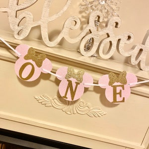 Op de afbeelding: Roze en gouden glitter Mickey Mouse-thema banner met het woord "ONE" in gouden letters. De banner hangt aan een witte schouw met een houten bord waarop "blessed" staat.
