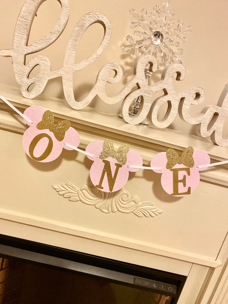 Op de afbeelding: Roze en gouden glitter Mickey Mouse-thema banner met het woord "ONE" in gouden letters. De banner hangt aan een witte schouw met een houten bord waarop "blessed" staat.