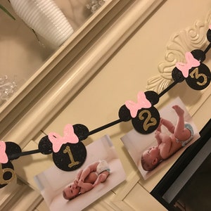May include: A black and pink Minnie Mouse-themed photo banner with gold numbers and pink bows. The banner displays photos of a baby, with numbers indicating months. The banner is a decorative item for a baby's milestone.
