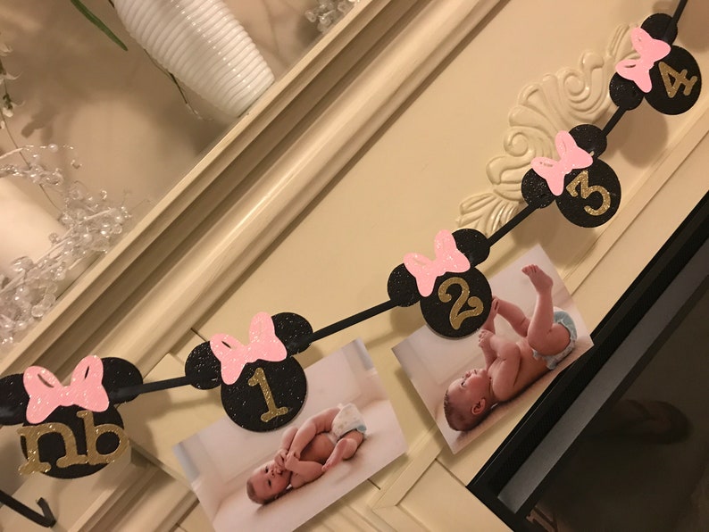 May include: A black and pink Minnie Mouse-themed photo banner with gold numbers and pink bows. The banner displays photos of a baby, with numbers indicating months. The banner is a decorative item for a baby's milestone.