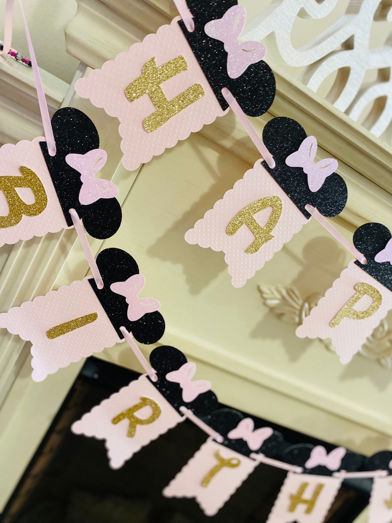 May include: Pink and black banner with gold glitter letters spelling "Happy Birthday". Each letter is attached to a black Mickey Mouse ear with a pink bow.