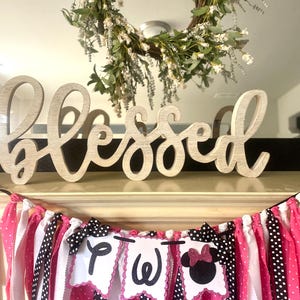 May include: A wooden "Blessed" sign sits above a banner with pink, white, and black fabric strips. The banner has three pennants with the letters "P", "W", and a Minnie Mouse silhouette. A floral wreath hangs above.