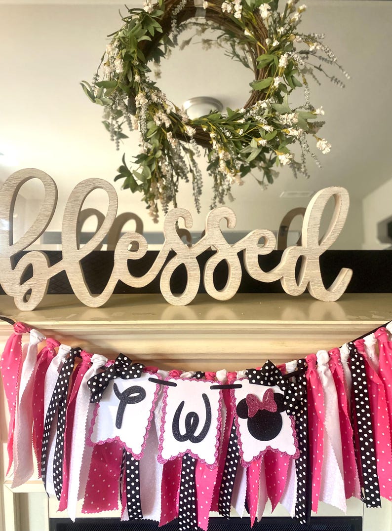 May include: A wooden "Blessed" sign sits above a banner with pink, white, and black fabric strips. The banner has three pennants with the letters "P", "W", and a Minnie Mouse silhouette. A floral wreath hangs above.