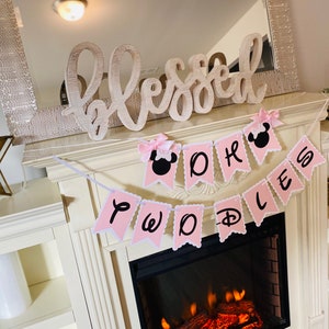 May include: Pink and white banner with the words "Two Worlds" and a Mickey Mouse head. The banner is hanging above a fireplace with a white mantel and a wooden sign that says "Blessed".