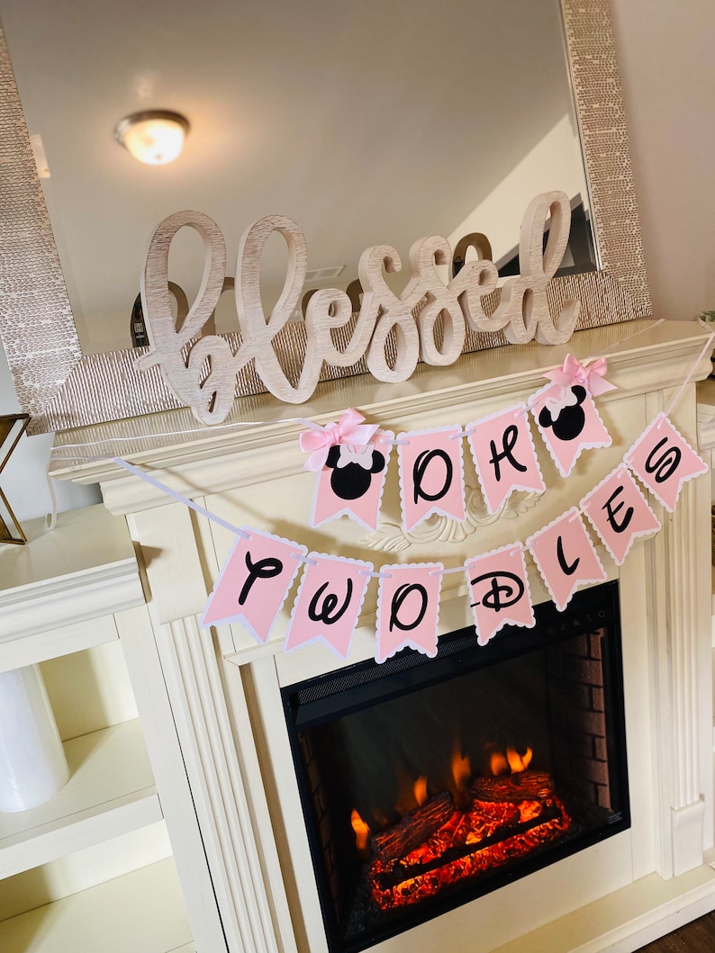 May include: Pink and white banner with the words "Two Worlds" and a Mickey Mouse head. The banner is hanging above a fireplace with a white mantel and a wooden sign that says "Blessed".