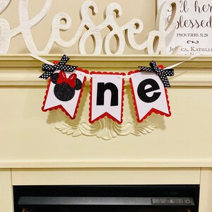 May include: A decorative banner spelling the word one, featuring three white pennants with red scalloped glitter borders. The first pennant displays a black glitter mouse silhouette with a red bow, while the others show black glitter letters.